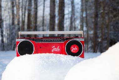Kuvassa on keskellä punainen matkaradio ja sen edessä on kaksi lumikumpua. Taustalla on metsää. 