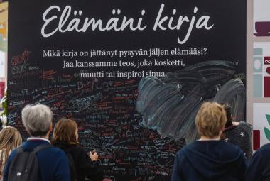 Kuvassa on seinämä, jossa lukee kirjoituskirjaimin Elämäni kirja ja sen alla pientä tekstiä mustalla pohjalla. Edessä on ihmisiä katselemassa seinää.