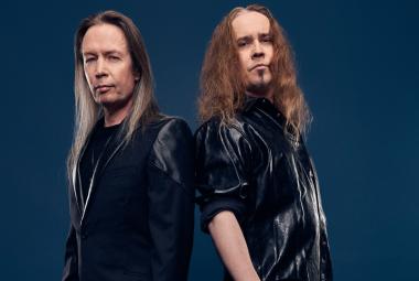 Kuvassa on vasemmalla Timo Kotipelto ja oikealla Jarkko Ahola. He ovat osittain selin ja molemmilla on mustat puserot ja pitkät hiukset.