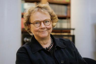 Kuvassa on Laura Kolbe puolivartalokuvassa istumassa ja hänellä on musta pusero päällään.  Taustalla on himmeästi kirjahyllyä.